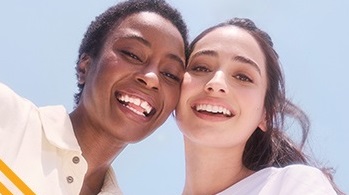 Eine Frau mit dunklem Hautton und eine Frau mit mittlerem Hautton stehen vor einem hellblauen Hintergrund. Beide Frauen lächeln und haben eine positive Ausstrahlung.