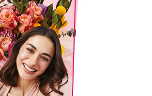 Eine Frau mit braunem Haar und strahlendem Teint lächelt in die Kamera. Im Hintergrund ist ein Blumenstrauß zu sehen.