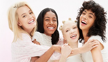 Vier fröhliche, junge Frauen mit unterschiedlichem Teint und Hauttyp umarmen sich und lächeln in die Kamera