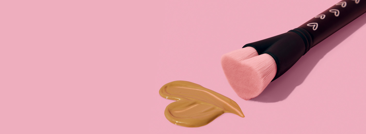 Der Mary Kay Heart-Shaped Foundation Brush – ein schwarzer Pinsel zum Auftragen von Make-up mit rosa Pinselhaaren in Herzform, für den Mary Kay bei jedem Verkauf an FLY & HELP spendet – zeichnet vor einem rosa Hintergrund ein Herz aus Make-up.