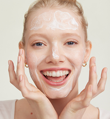 Eine Frau mit strahlendem Lächeln trägt mit beiden Händen ein Peeling im gesamten Gesicht auf