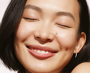 Makroaufnahme einer brünetten asiatischen Frau, die mit geschlossenen Augen lächelt, umgeben von Sonnenlicht.