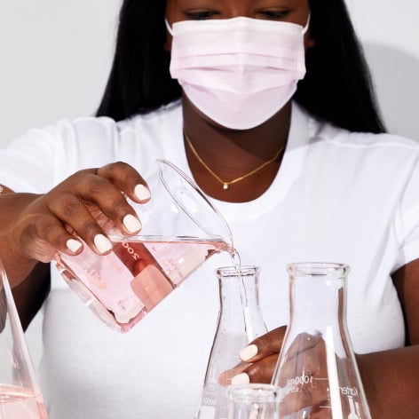 In einem Labor kippt eine Frau, die eine Maske trägt, eine rosa Flüssigkeit von einem in ein anderes Glas