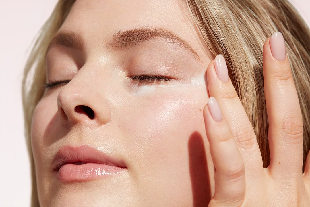 Junge Frau mit blonden Haaren trägt mit geschlossenen Augen Augencreme auf ihre Haut auf