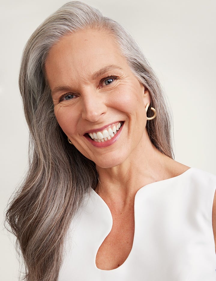 Eine Frau in ihren 60ern mit reifer Haut, langem silbernen Haar und einem gepflegten Hauterscheinungsbild lächelt und blickt in die Kamera