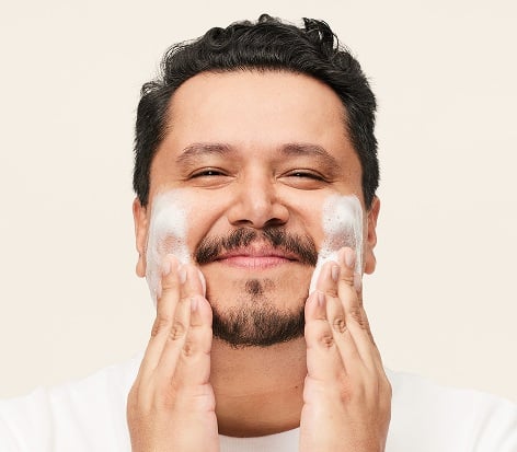 Ein Mann bei der Hautpflege-Routine: Er trägt mit beiden Händen Cleanser auf den Wangen auf und reinigt sein Gesicht