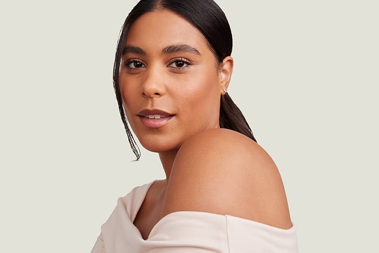 Junge Frau mit glatter Haut und dezentem Make-up blickt selbstbewusst in die Kamera, trägt ein schulterfreies, cremefarbenes Oberteil