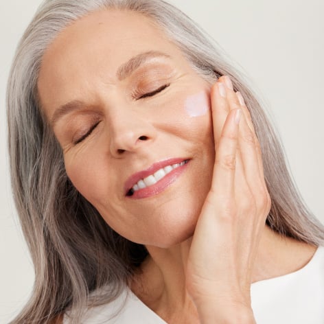 Eine Frau mit reifer, strahlender Haut und silbernem Haar hat die Augen geschlossen und trägt ein Serum in ihrem Gesicht auf