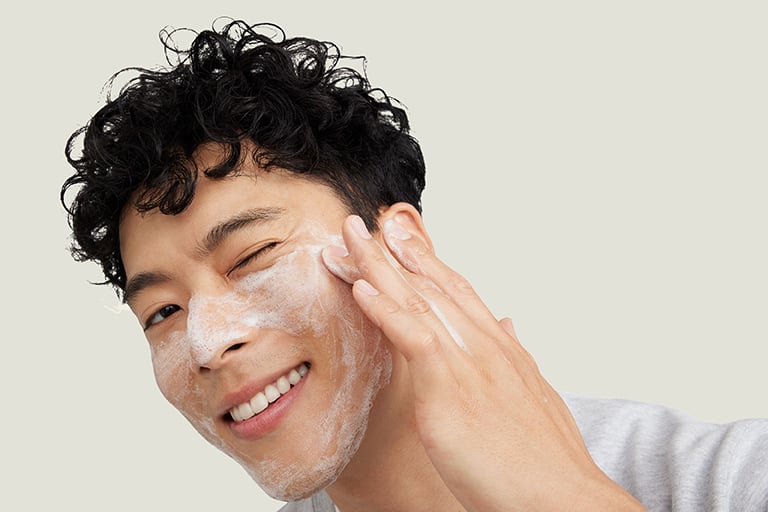 Junger Mann mit aufgeschäumtem Gesichtsreiniger auf der Haut, der charmant zur Seite in die Kamera zwinkert