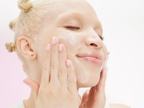 Eine junge Frau mit heller Haut und blondem Haar trägt mit geschlossenen Augen ein Peeling im Gesicht auf