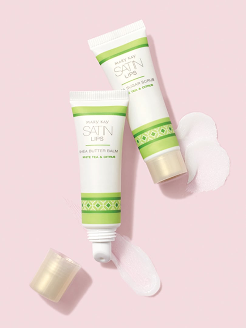 Mary Kay Satin Lips Set, das aus Shea Sugar Scrub und Shea Butter Balm besteht mit Produktklecksen vor einem rosanen Hintergrund