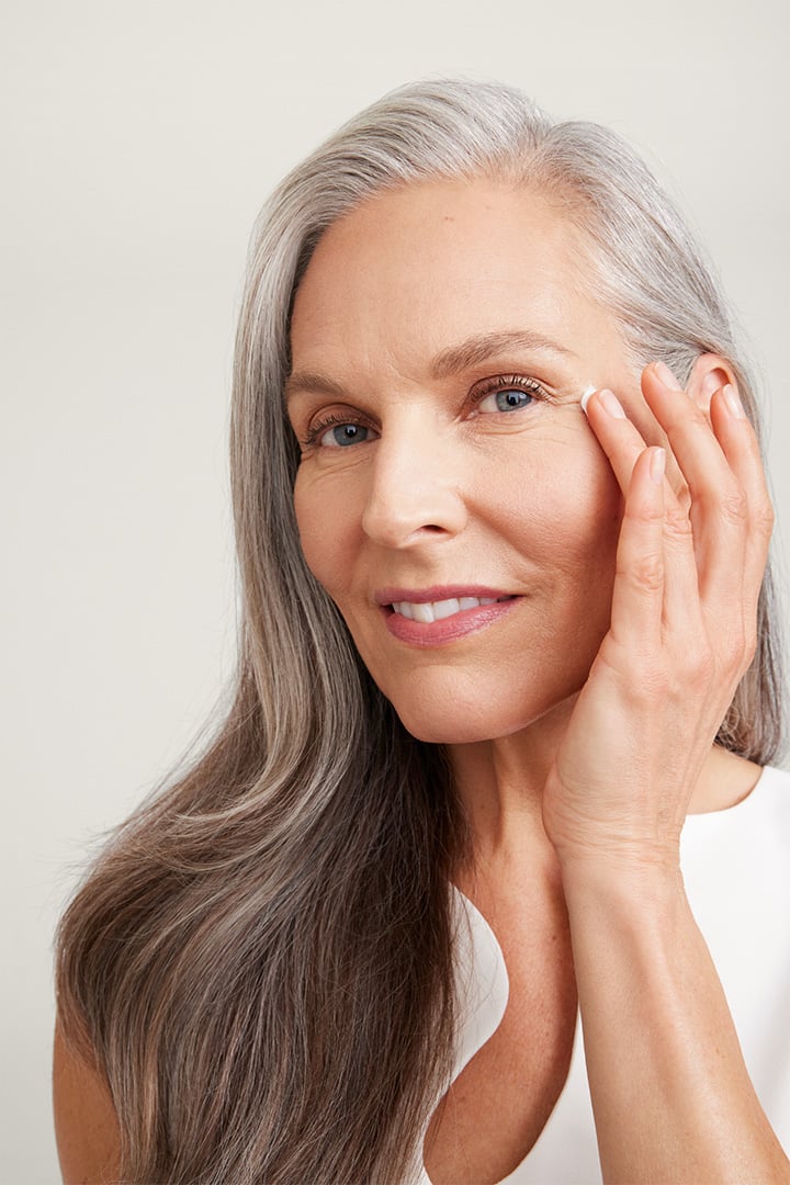 Eine Frau in ihren 60ern mit reifer Haut, langem silbernen Haar und einem gepflegten Hauterscheinungsbild lächelt in die Kamera während sie mit dem Finger Augencreme aufträgt