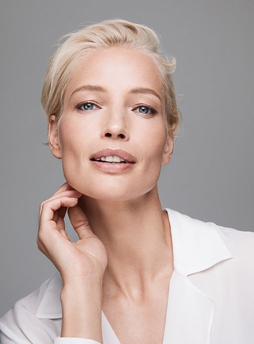 Eine Frau in den 40ern mit hellblondem Haar und gepflegtem Teint blickt in die Kamera und berüht mit ihrer Hand ihren Hals