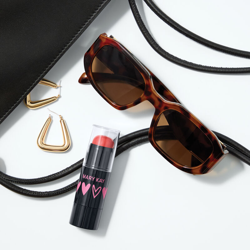 Limited-edition Mary Kay Blush Stick in Kind Spirit shade with tortoiseshell sunglasses, gold hoop earrings, a black clutch on a white background.
