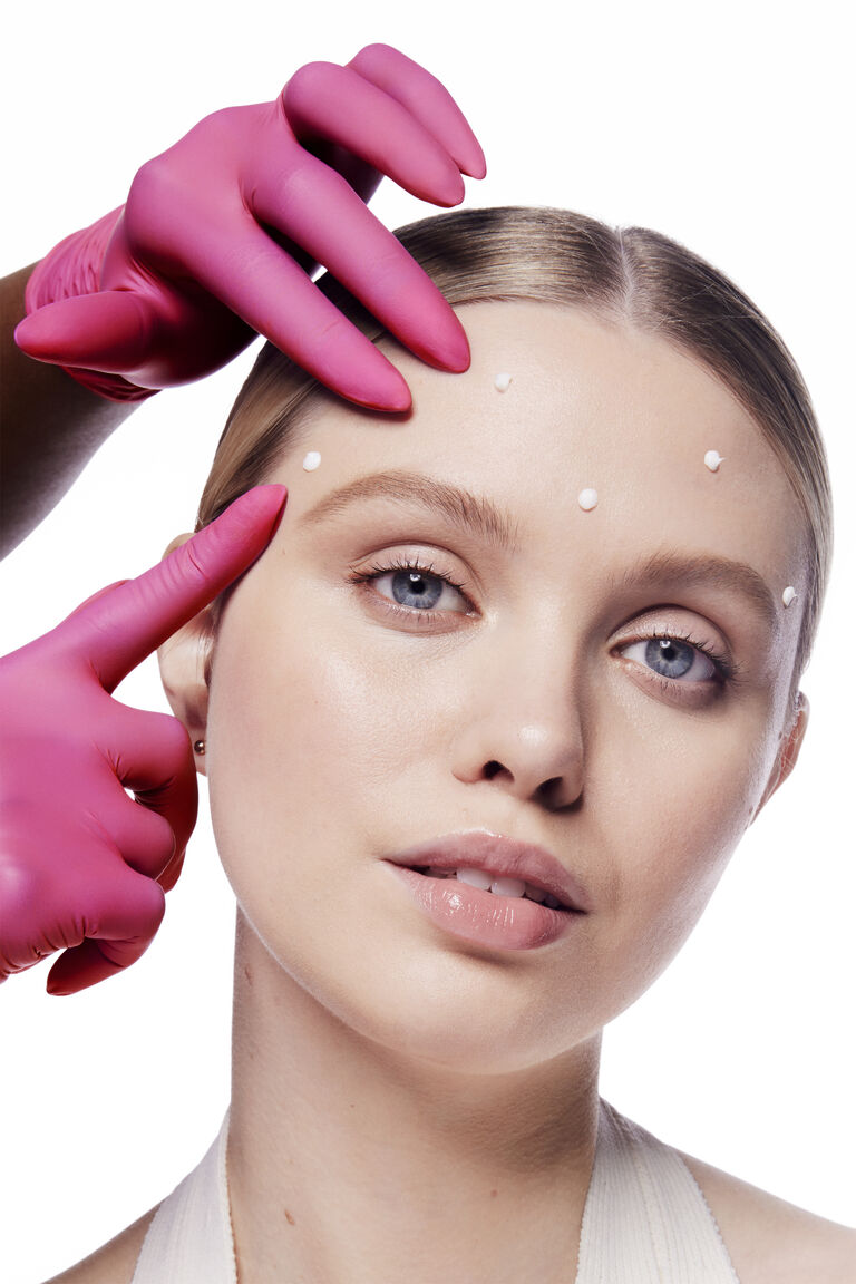 Eine Frau mit Tupfern des &bdquo;Mary Kay Dynamic Wrinkle Limiter&ldquo; auf der Stirn. Eine Hand mit rosa Handschuh ber&uuml;hrt ihre Stirn, um einen wissenschaftlichen Effekt zu erzeugen.