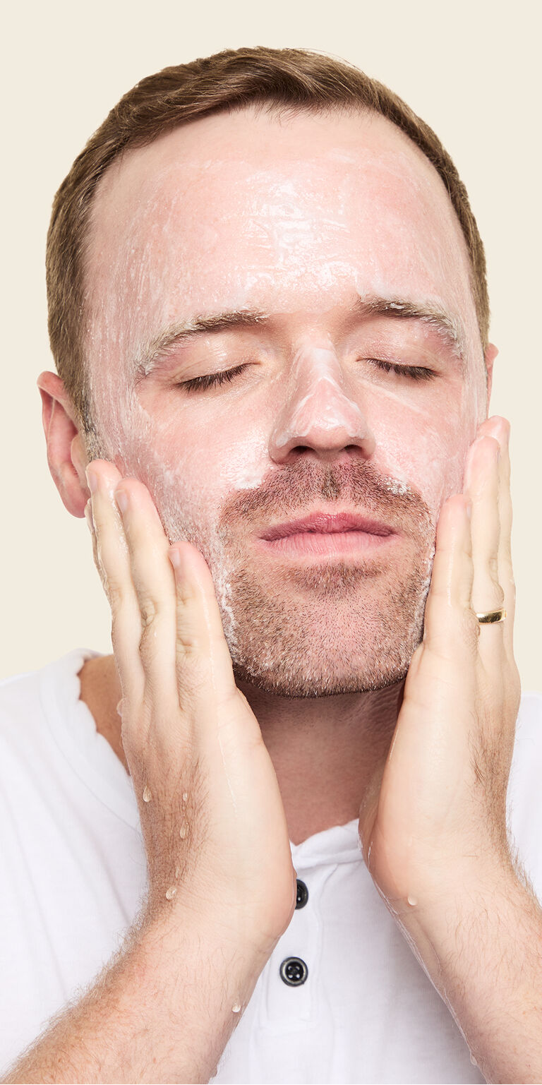 Ein Mann tr&auml;gt sich mit beiden H&auml;nden und geschlossenen Augen das MK Men Invigorating Face Scrub im Gesicht auf und massiert es ein (Peeling)