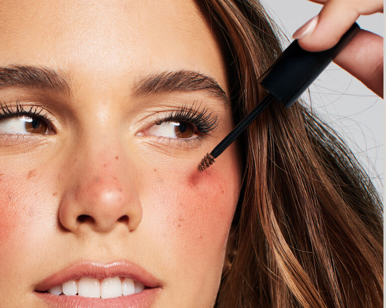 Eine Frau, deren Gesicht mit dem Strawberry Makeup Trend geschminkt ist: Fake Freckles, glowy skin und sonnengek&uuml;sste Wangen. Mit einer Hand tr&auml;gt sie weitere Fake Freckles auf.