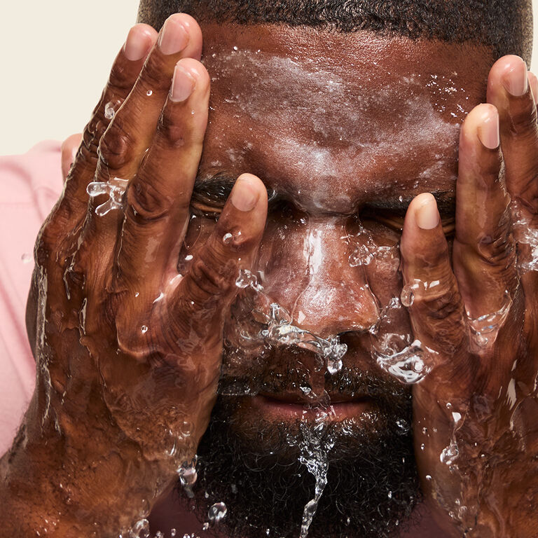 Nahaufnahme eines Mannes, der sich mit beiden H&auml;nden das MK Men Invigorating Face Scrub (Gesichtspeeling) mit Wasser aus dem Gesicht sp&uuml;lt