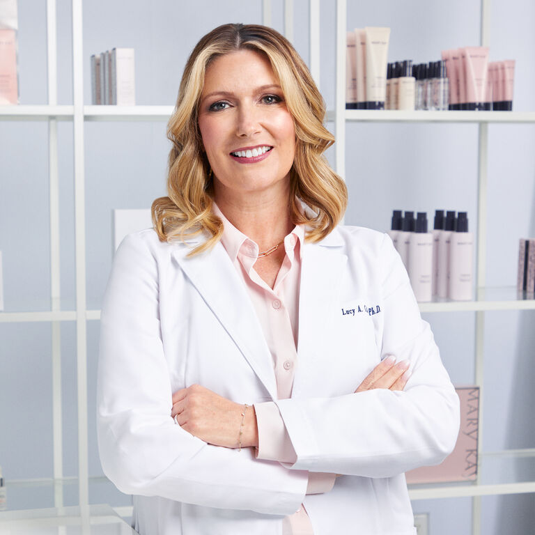 Dr. Lucy Gildea, Chief Brand and Scientific Officer bei Mary Kay, tr&auml;gt einen Laborkittel. Hinter ihr im Regal sind Mary Kay Gesichtspflegeprodukte sichtbar.