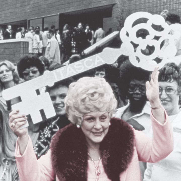 Mary Kay Ash, die Firmengr&uuml;nderin des Direktvertriebsunternehmens Mary Kay, ist auf einem alten Foto im Vordergrund in einem rosa Kost&uuml;m zu sehen. Sie h&auml;lt mit beiden H&auml;nden einen gro&szlig;en Schl&uuml;ssel in die H&ouml;he. Hinter ihr sind viele weitere Menschen zu sehen.