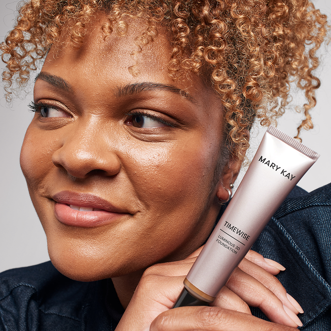 Eine Frau mit lockigem Haar h&auml;lt die Mary Kay TimeWise&reg; Luminous 3D Foundation neben dem Gesicht und pr&auml;sentiert das Produkt.
