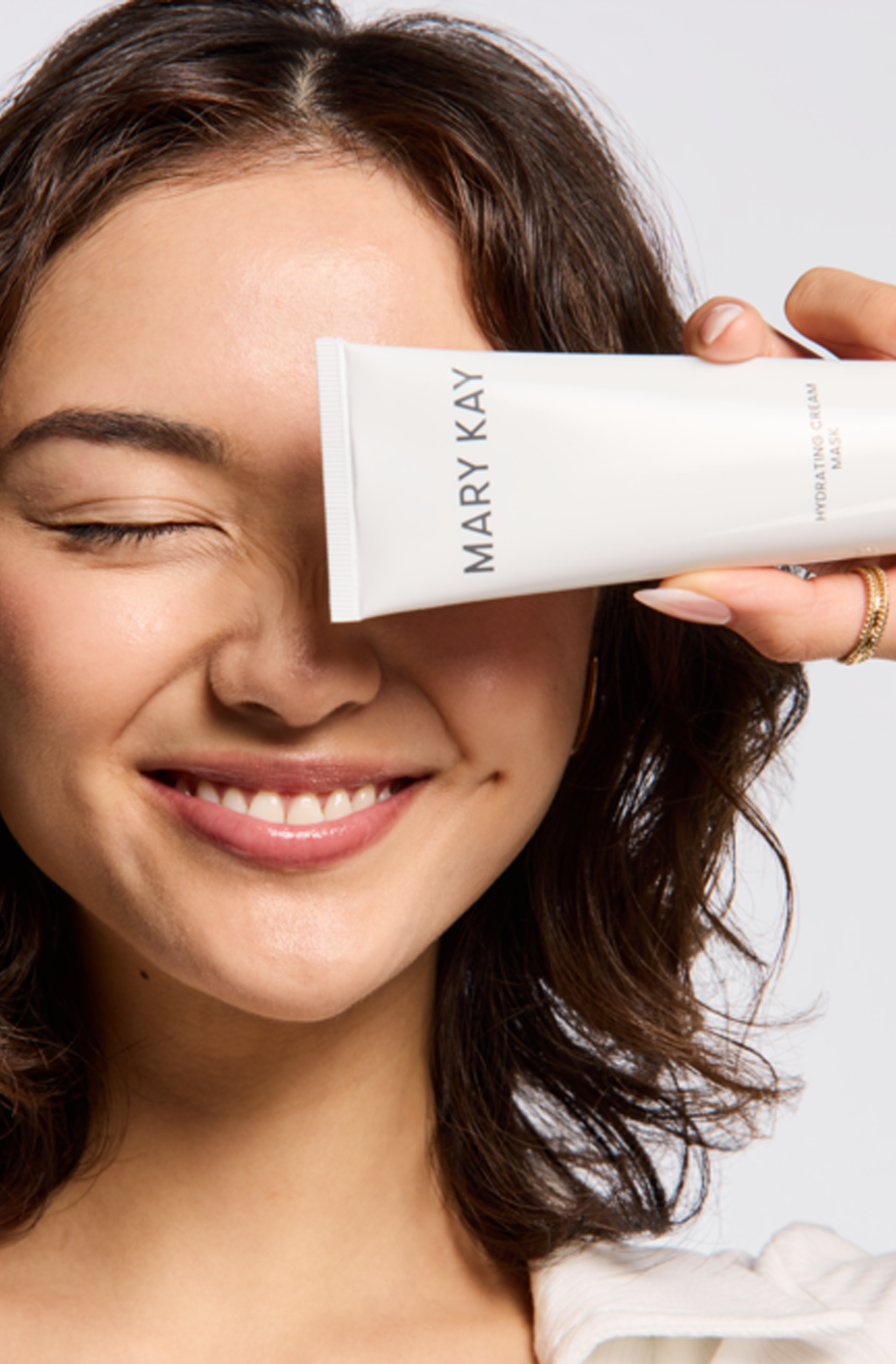 Eine lachende Frau mit geschlossenen Augen hält eine Tube der Hydrating Cream Mask vor ihr Gesicht.