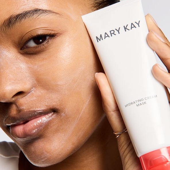 Nahaufnahme einer Frau, in deren Gesicht eine d&uuml;nne Schicht der Mary Kay Hydrating Cream Mask aufgetragen ist. Mit ihrer Hand h&auml;lt die Frau die Tube dieser Leave-on Gesichtsmaske neben ihr Gesicht.